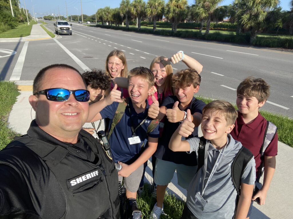 A group of middle-grade kids pose and smile for a group selfie with a law enforcement officer.