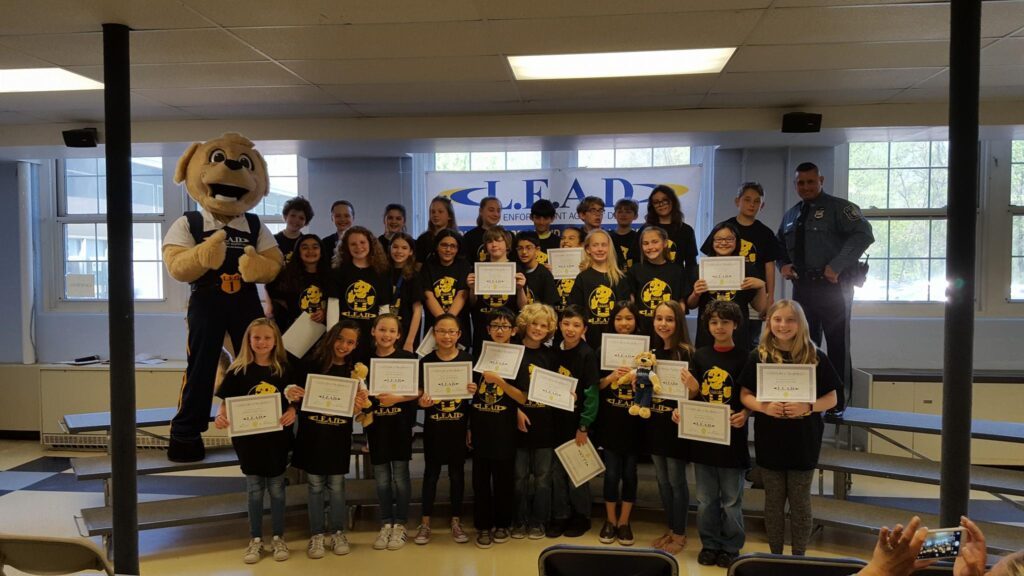 A large class of students from Harding Township pose with certificates and matching t-shirts accompanied by a police officer and L.E.A.D. mascot dog.